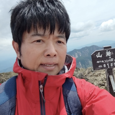 山頂の標識の前に立つ、赤いジャケット姿の平貞さん